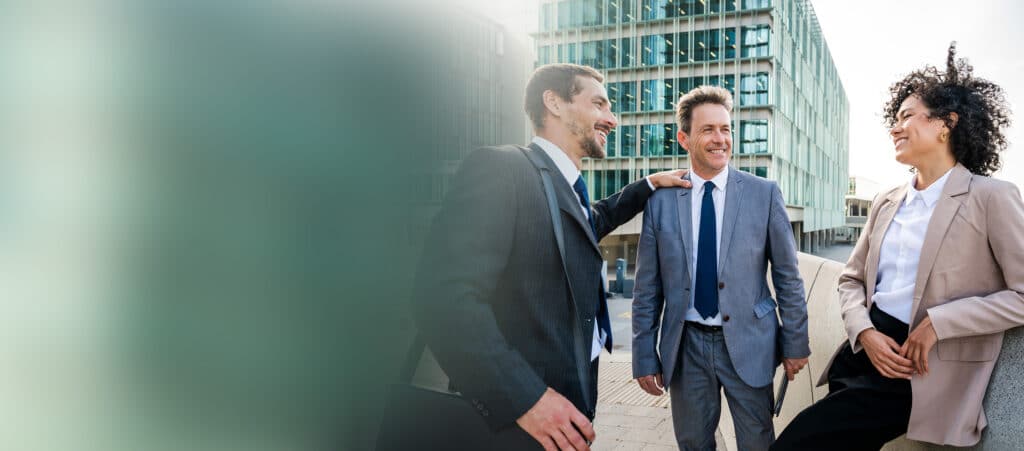 Drei Geschäftsleute, zwei Männer und eine Frau, stehen im Freien vor einem modernen Bürogebäude, lächeln und diskutieren über ein Nutzungsdauer-Gutachten, während sie in formeller Geschäftskleidung gekleidet sind.